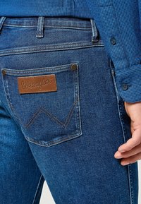 Jeans de denim azul con un parche de cuero marrón de Wrangler en el bolsillo trasero. La tela tiene una textura clásica con un leve desgaste y detalles de costura.