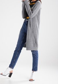 Grå stickad cardigan med långa ärmar, bärs över en randig topp; matchad med högmidjade blå jeans och vita klacksandaler.