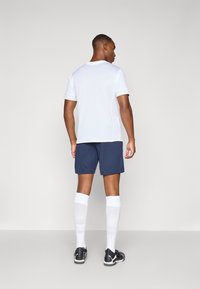 T-shirt de sport blanc à manches courtes, shorts navy, chaussettes blanches montantes et chaussures de sport noires. Positionné de dos sur un fond clair.