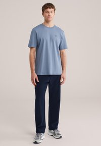 Jonge man die recht vooruit kijkt, gekleed in een lichtblauw T-shirt met korte mouwen, donkerblauwe broek en grijze sportschoenen, voor een effen achtergrond.