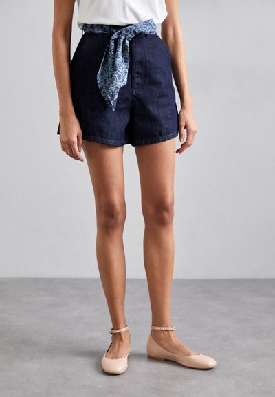 Șorturi din denim de culoare albastru închis, cu talie înaltă și buzunare laterale, asortate cu o eșarfă modelată albastră legată la talie și balerini nud.