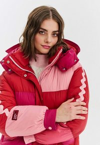 Žena s ramenními vlasy hnědé barvy, oblečená v zateplené bundě s vysokým límcem v červené a růžové barvě, se zkříženýma rukama, dívající se do kamery.