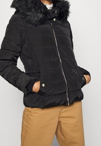 Černá péřová bunda s umělou kožešinou na kapuci, zipem vpředu, bočními kapsami a prošitým texturem. Obsahuje elastické manžety a pas.