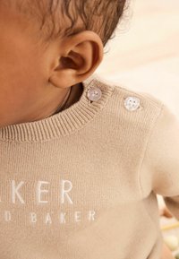 Pull en tricot beige clair avec col côtelé, comportant un texte brodé blanc et des boutons décoratifs à l'épaule.