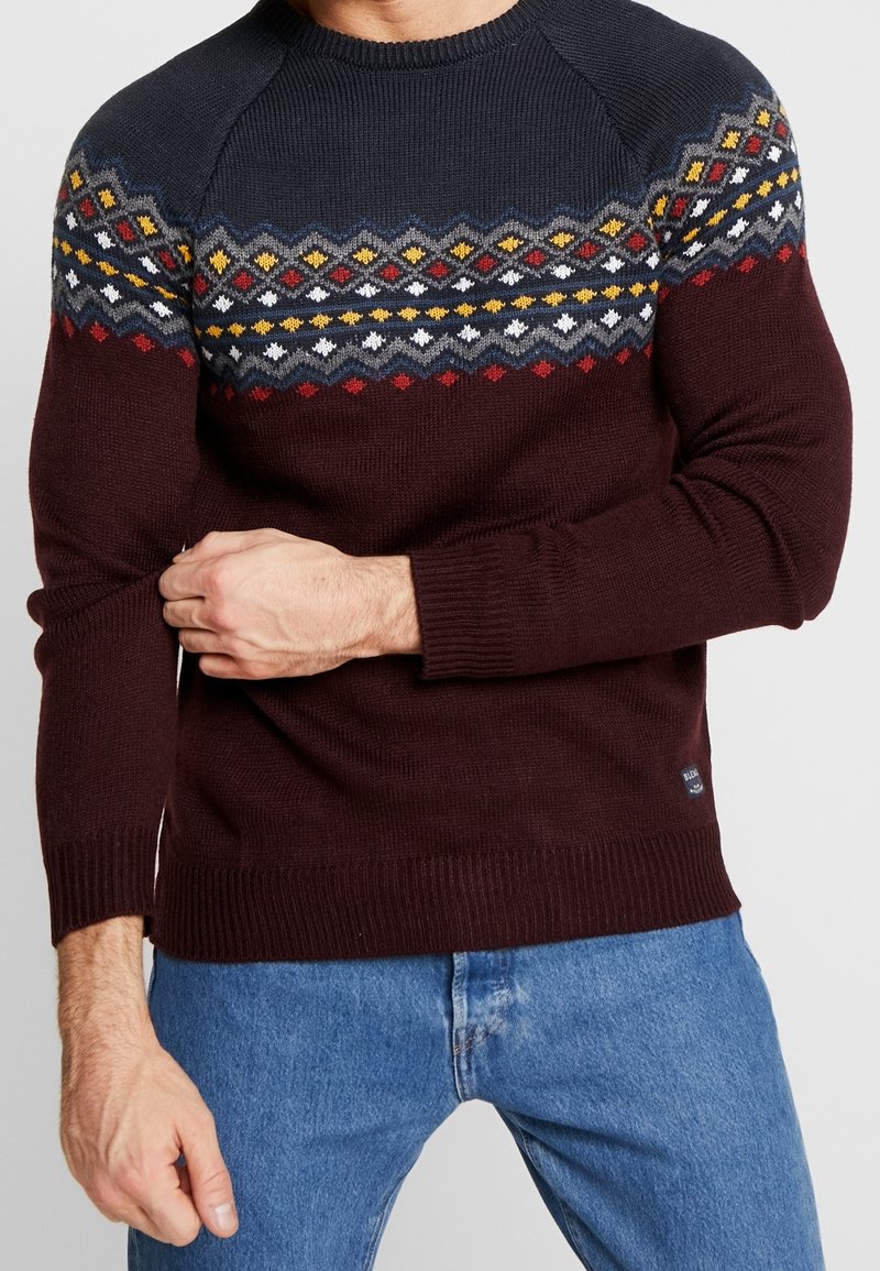 Pull en maille bordeaux avec un empiècement à motif orné d'accents bleus, jaunes et rouges. Poignets et ourlet côtelés, col rond.