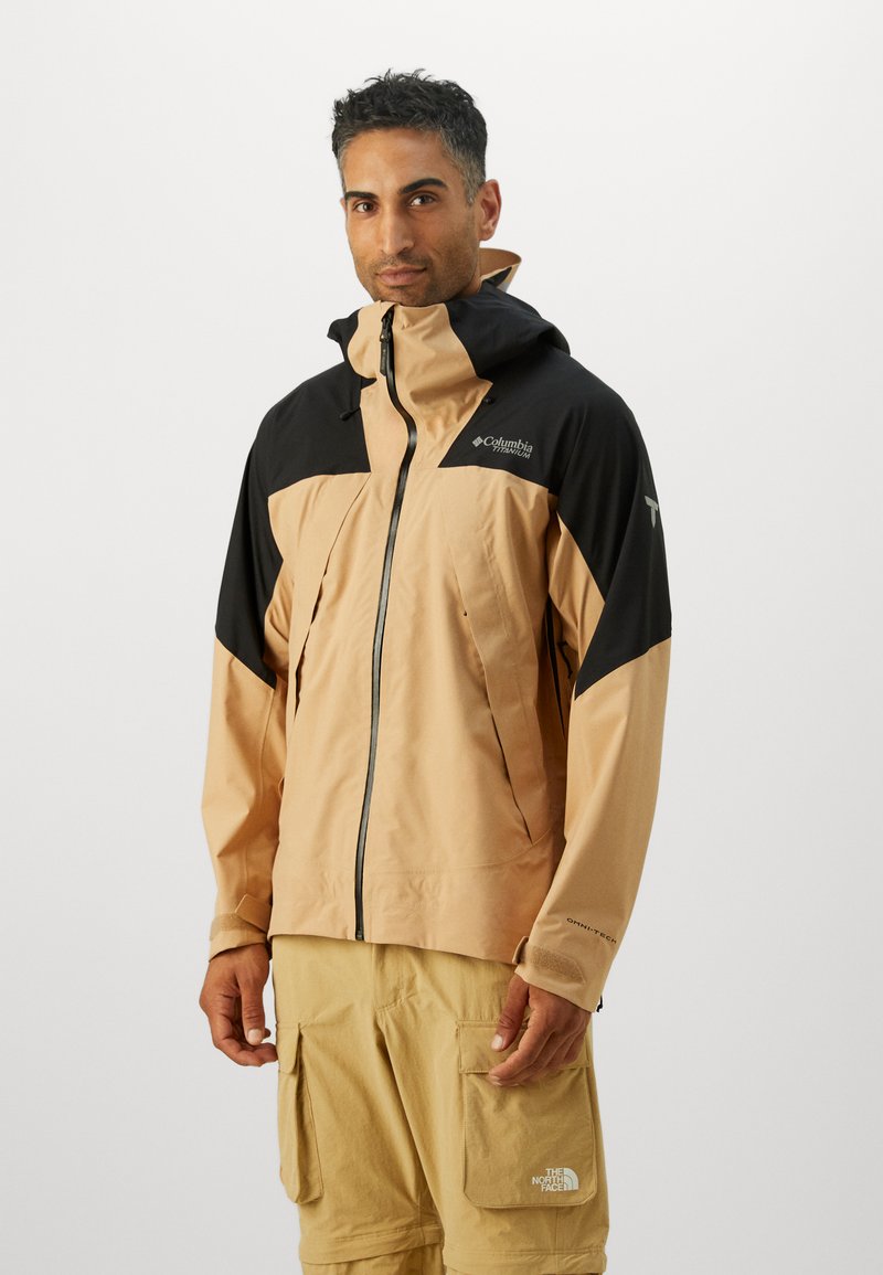 La veste imperméable présente un corps beige avec des accents noirs, un col haut, une capuche ajustable et une fermeture à glissière à l'avant. Tissu durable avec une finition texturée.