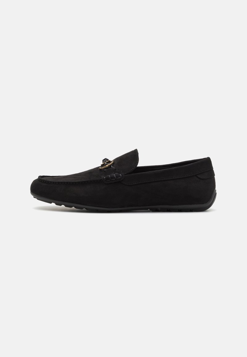 Mocassin en daim noir avec semelle en caoutchouc basse et détail de boucle en métal doré sur la partie supérieure, présenté de profil sur fond blanc.