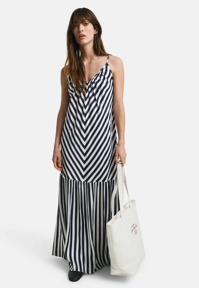 Trägerloses Maxikleid mit marineblauen und weißen Zickzackstreifen, gerafftem Oberteil und plissiertem Saum. Kombiniert mit einer cremefarbenen Tragetasche.
