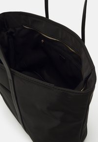 Sac cabas noir avec longues anses, ouverture supérieure montrant une poche intérieure zippée avec fermeture éclair dorée sur fond blanc.
