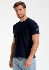 T-shirt a maniche corte blu navy con superficie texturizzata, scollatura rotonda e una patch con il marchio sulla manica sinistra. Abbinato a jeans azzurri.