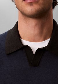 Άνδρας που φοράει σκούνο μπλε πουλόβερ με μαύρο γιακά πάνω από λευκό μπλουζάκι με στρογγυλή λαιμόκοψη, δείχνοντας τον λαιμό και το κάτω μέρος του προσώπου.