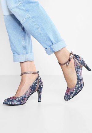 Fødder iført blå højhælede sko med blomsterstof og ankelsnore, kombineret med lyseblå opbukkelige jeans mod en hvid baggrund.