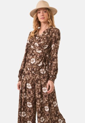 Mujer con vestido tipo wrap de manga larga en teñido anudado marrón y blanco, y sombrero beige de ala ancha, posando frente a un fondo liso.