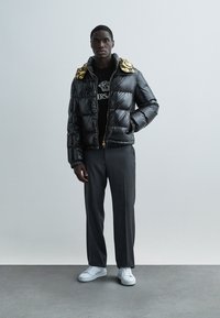 Musta kiiltävä toppatakki, jossa on kullanvärisillä yksityiskohdilla varustettu huppu, yhdistetty mustaan graafiseen T-paitaan ja harmaisiin housuihin. Valkoiset tennarit viimeistelevät asun.