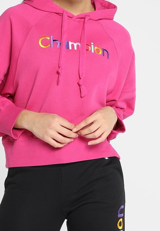 Personne portant un sweat à capuche rose vif avec un texte "Champion" multicolore et un pantalon noir avec une partie du logo visible sur la jambe.