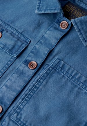 Niebieska koszula dżinsowa z dwoma przednimi kieszeniami, brązowymi guzikami i kołnierzykiem. Materiał ma teksturowane wykończenie z delikatnym przetarciem.