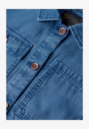 Blaue Jeanshemd mit zwei Vordertaschen, braunen Knöpfen und einem Kragen. Der Stoff zeigt eine strukturierte Oberfläche mit subtiler Verwaschung.