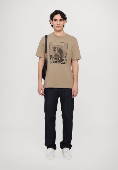 T-shirt en coton beige avec une impression graphique portant la mention "TE SOUVIENS-TU DE L'ENNUI ?" associée à un jean en denim foncé et des baskets blanches. Sac à bandoulière noir.