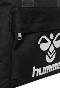 Svart nylonväska med en framficka med dragkedja, stor vit logotyp och texturerad yta. Inkluderar robusta bärremmar.