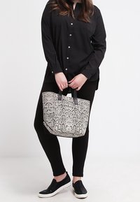 Czarna koszula zapinana na guziki, czarne legginsy i czarne buty wsuwane. Torba na ramię ma szary i czarny wzór w węża oraz solidne uchwyty.