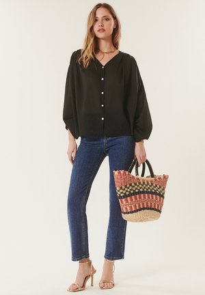Femme en blouse noire boutonnée, jean bleu et sandales à talons hauts tenant un sac cabas tissé multicolore sur un fond uni.