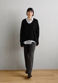 Zwarte wolmix oversized sweater, met V-hals, gedragen over een wit en zwart gestreept overhemd met knopen; gecombineerd met grijze ribfluwelen broek en zwarte schoenen.