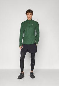 Žalias ilgomis rankovėmis sportinis topas su ketvirčio užtrauktuku ir pilkomis detalėmis, derinamas su juodomis šortais ir prigludusiomis juodomis leggings, papildytas juodais sportiniais bateliais.