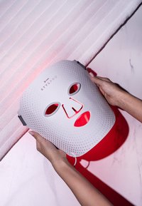 Vit LED-ansiktsmask med perforerat mönster, röda detaljer och urskurna ögon- och munöppningar. Hålls av två händer mot en texturerad bakgrund.