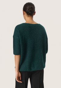 Oversized groene gebreide trui met korte mouwen en een brede halslijn, met een gestructureerde afwerking en een ontspannen pasvorm, in combinatie met zwarte broek.