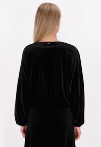 Sort velour top med løs pasform, ballonermer og indsnævret talje. Har et lille metallisk mærke på den bagerste halsudskæring.