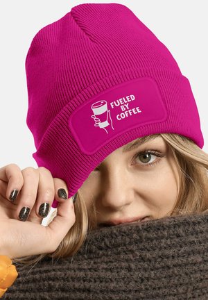Fuchsia knit beanie with a patch featuring the text "FUELED BY COFFEE" and an illustration of a coffee cup, worn casually.