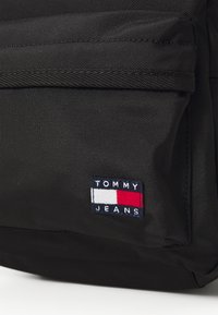 Sac à dos en tissu noir avec une surface texturée, une poche avant et un patch logo brodé avec des accents bleu, blanc et rouge.