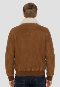 Veste bomber en daim marron avec un col en shearling crème, un ourlet côtelé et un détail de couture sur le yoke arrière. Portée avec un jean en denim foncé.