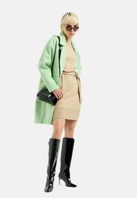 Cappotto oversize verde menta, vestito lavorato a maglia beige con cintura, stivali neri in vernice fino al ginocchio e occhiali da sole, caratterizzati da texture lisce e forme strutturate.
