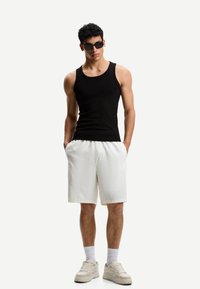 Cămașă neagră cu dungi, pantaloni scurți albi, adidași bej și ochelari de soare; modelul stă cu mâinile în buzunare pe un fundal deschis.