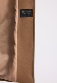 Wnętrze brązowego wełnianego płaszcza z czarną etykietą "Wool Collection Makalu Made in Poland" na jednolitym jasnym tle.