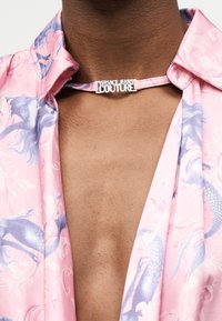 Κοντινό πλάνο ενός ατόμου που φοράει ροζ και μωβ πουκαμίσα με ανοιχτό γιακά και λευκό κολιέ τύπου «Versace Jeans Couture».