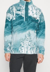 Pull à col demi-fermeture éclair en polaire bleu sarcelle et blanc, avec un motif de paysage montagneux et une poche zippée à l'avant. Logo Columbia présent.