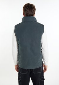 Dunkelgraue Pufferweste mit hohem Kragen, horizontalen Nähten und glatter Oberfläche, getragen über einem weißen Langarmshirt.