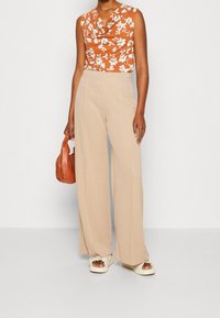 Blommig topp med cowl-hals i rostorange, tillsammans med beige vida byxor. Har en mjuk textur och en strukturerad silhuett. Håller en brun handväska.