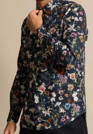 Homme ajustant le col d'une chemise à manches longues sombre avec un imprimé floral coloré et des boutons blancs, debout devant un fond beige uni.