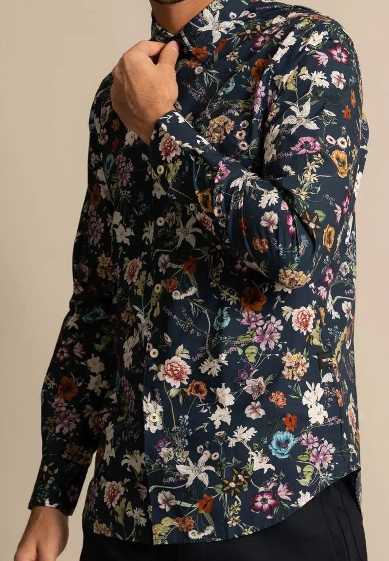 Homme ajustant le col d'une chemise à manches longues sombre avec un imprimé floral coloré et des boutons blancs, debout devant un fond beige uni.