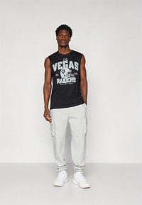 Camisola preta sem mangas com gráfico "Las Vegas Raiders", combinada com calças cargo cinza claro com bolsos laterais, usada com ténis brancos.