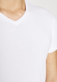 Close-up van een persoon die een effen wit V-hals T-shirt met korte mouwen draagt, waarbij de nek, schouder en het bovenste borstgebied zichtbaar zijn.