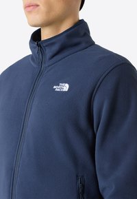Giacca in fleece blu navy con colletto alto, zip completa e tasche laterali con zip. Presenta un logo bianco sul petto e una superficie morbida e текстurizzata.