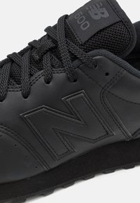 Chaussure de sport noire avec une tige en cuir, une languette en mesh, des lacets texturés et un logo "N" bien en vue. Détails de rembourrage sur la semelle.