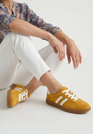 Person sitzt mit überkreuzten Beinen, trägt senfgelbe Sneakers mit weißen Streifen, weiße Hose und ein langärmeliges, geblümtes Hemd.