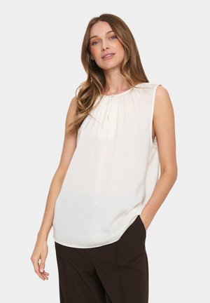 Blouse blanche sans manches avec un décolleté plissé et des détails de boutons, fabriquée en tissu léger, associée à un pantalon sombre.