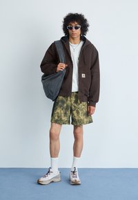 Felpa marrone con zip, t-shirt grigia, pantaloni corti mimetici, scarpe da ginnastica bianche e occhiali da sole. Porta una borsa a tracolla grigia con una texture morbida.