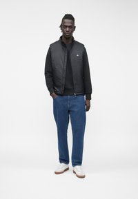 Schwarze gesteppte Weste über einem schwarzen Sweatshirt, kombiniert mit blauen Jeans und weißen Turnschuhen. Modell steht vor einem schlichten weißen Hintergrund.